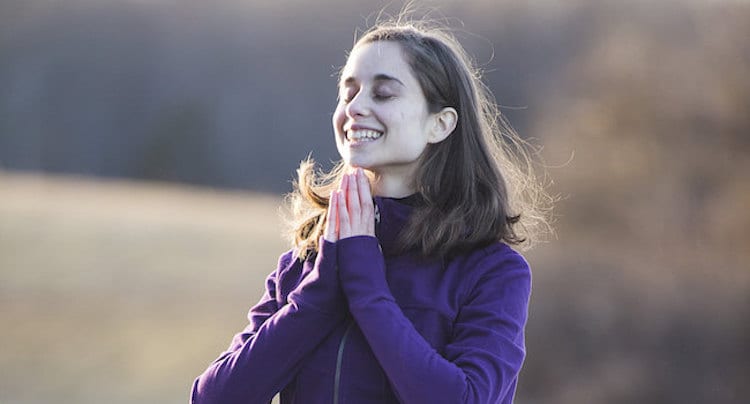 Woman with hands in prayer position, smiling, eyes closed, wearing a long sleeved purple shirt. 