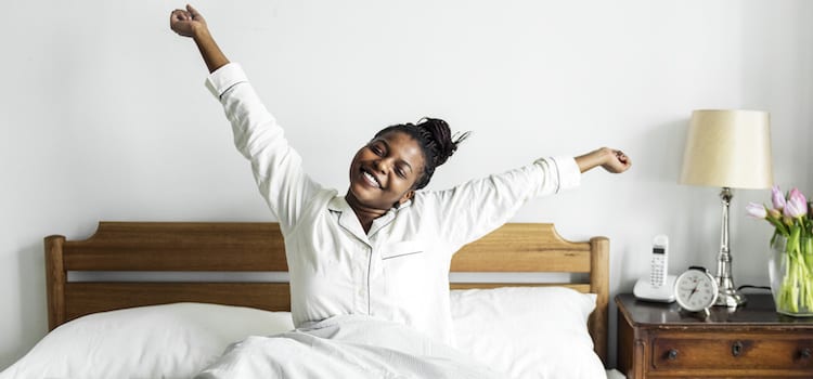 Woman waking up, siting in bed with arms stretched up smiling. 