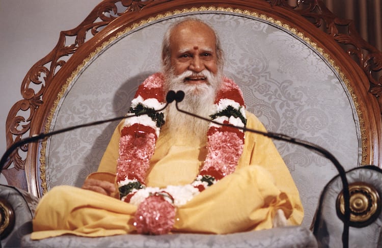 Swami Satchidananda sitting with a garland, smiling and talking into a microphone.